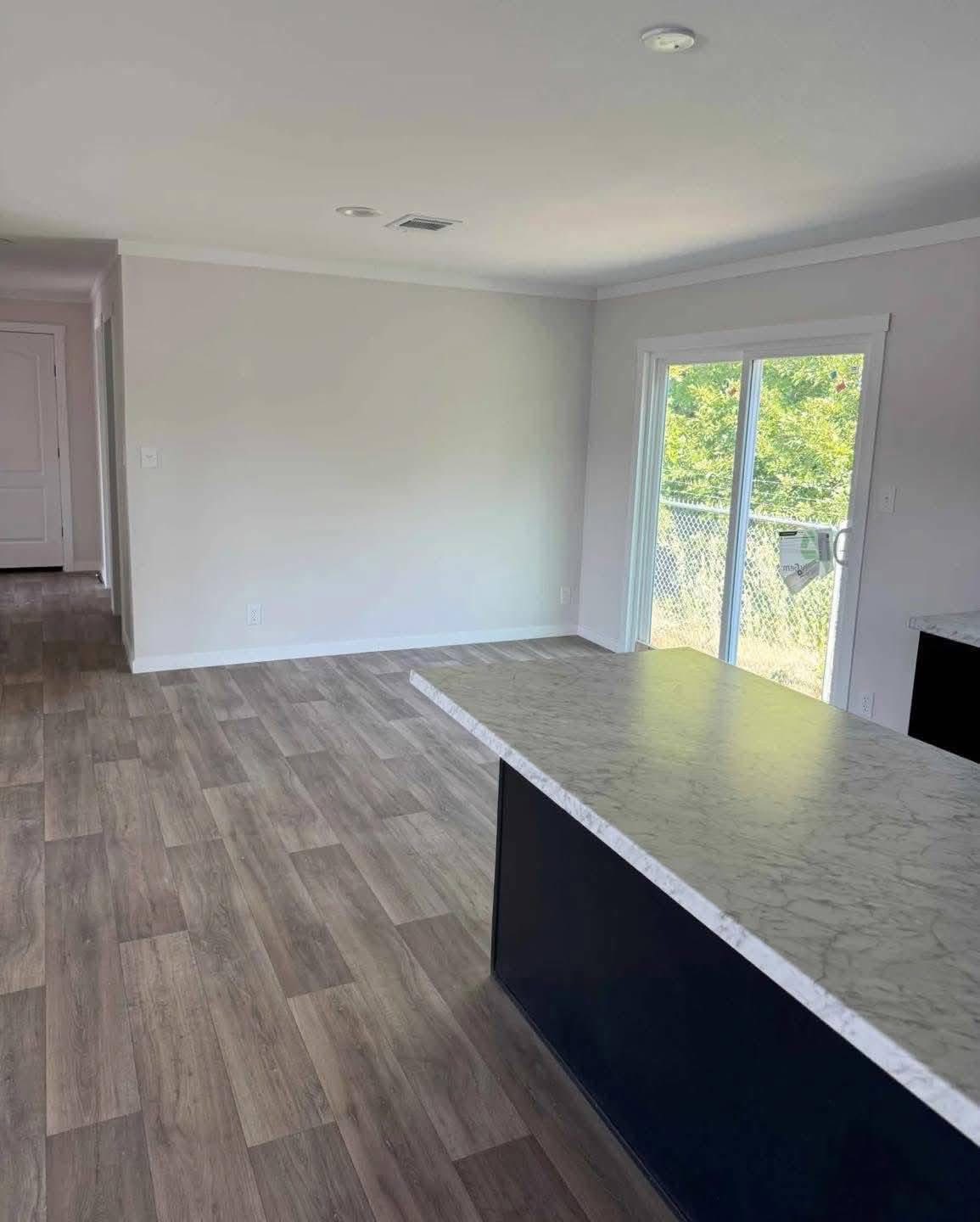 Modern open-plan room with light wood floors, white walls, and a marble countertop. Sliding glass door leads to a green outdoor view, creating a fresh and airy ambiance.