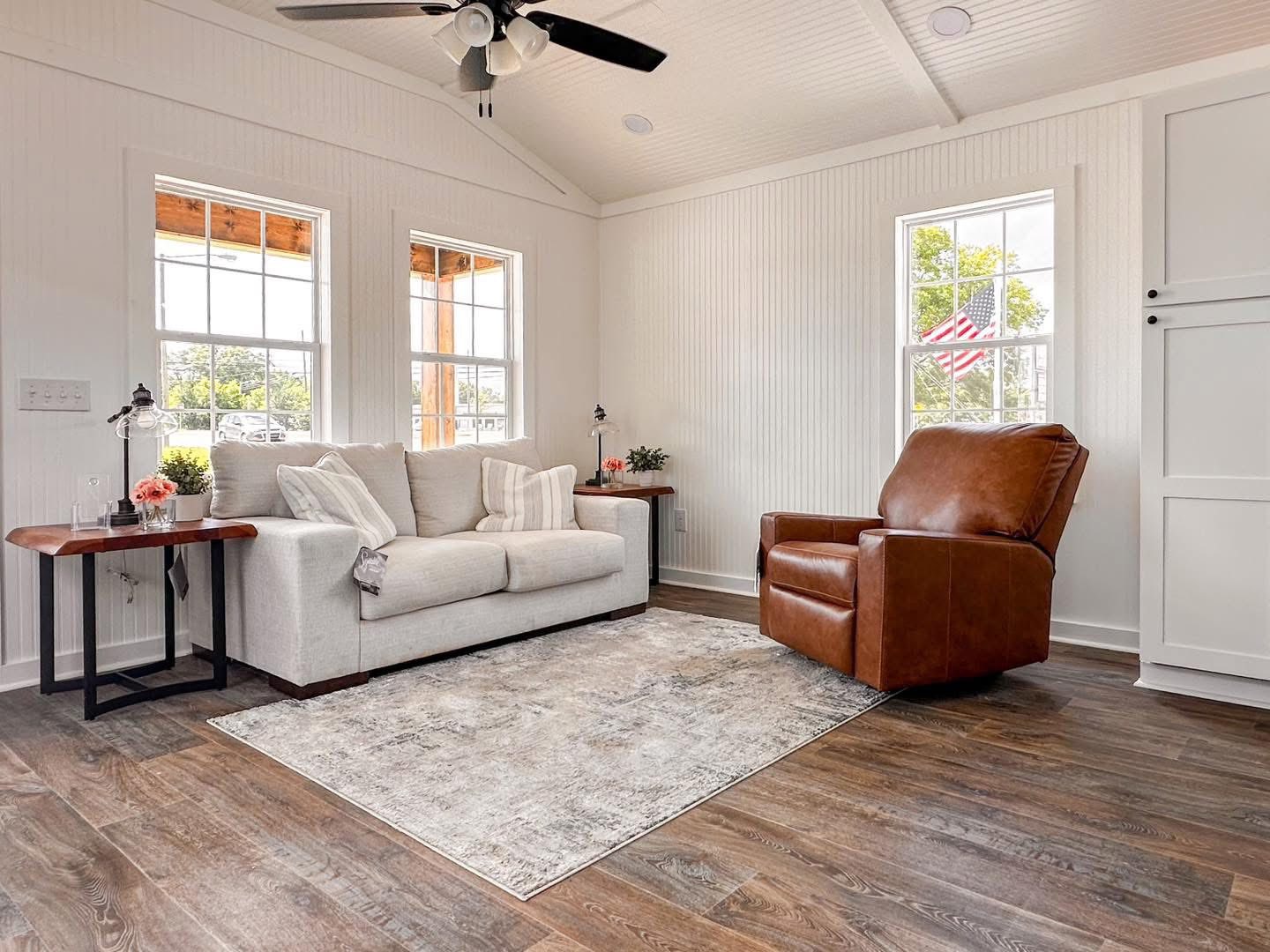 A cozy living room with a gray sofa and cushions, a brown leather armchair, and a gray rug on wooden flooring. Bright windows enhance the airy feel.