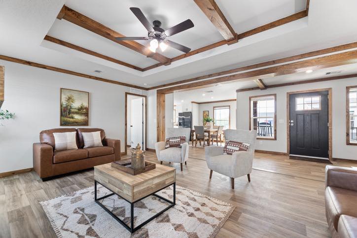 Bright living room with wood beams, ceiling fan, and wooden floor. Features a brown leather sofa, two gray chairs, a geometric rug, and a wooden coffee table.