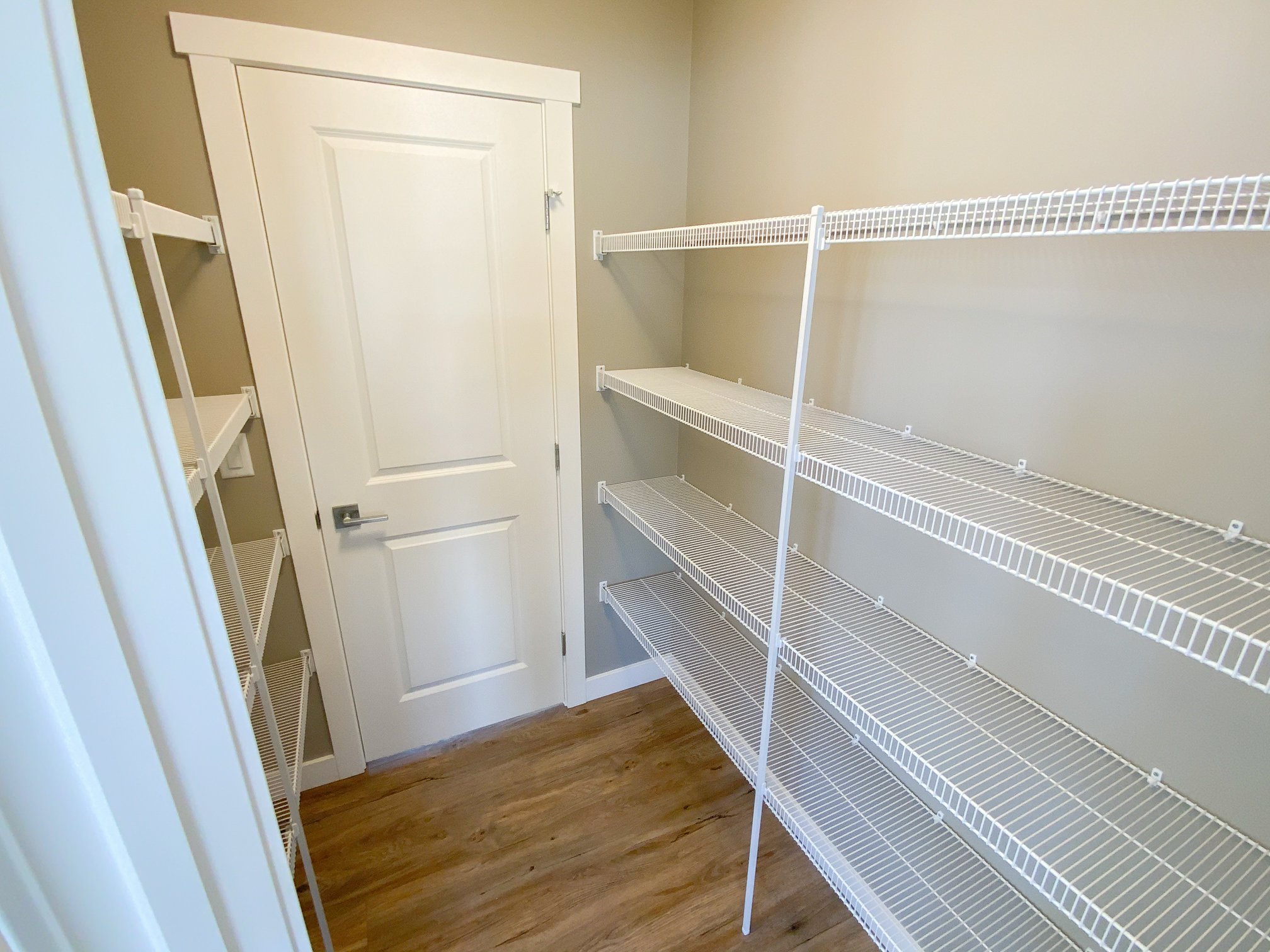 Small pantry with beige walls and wooden floor, featuring white wire shelving on both sides. A closed white door is at the end of the room.