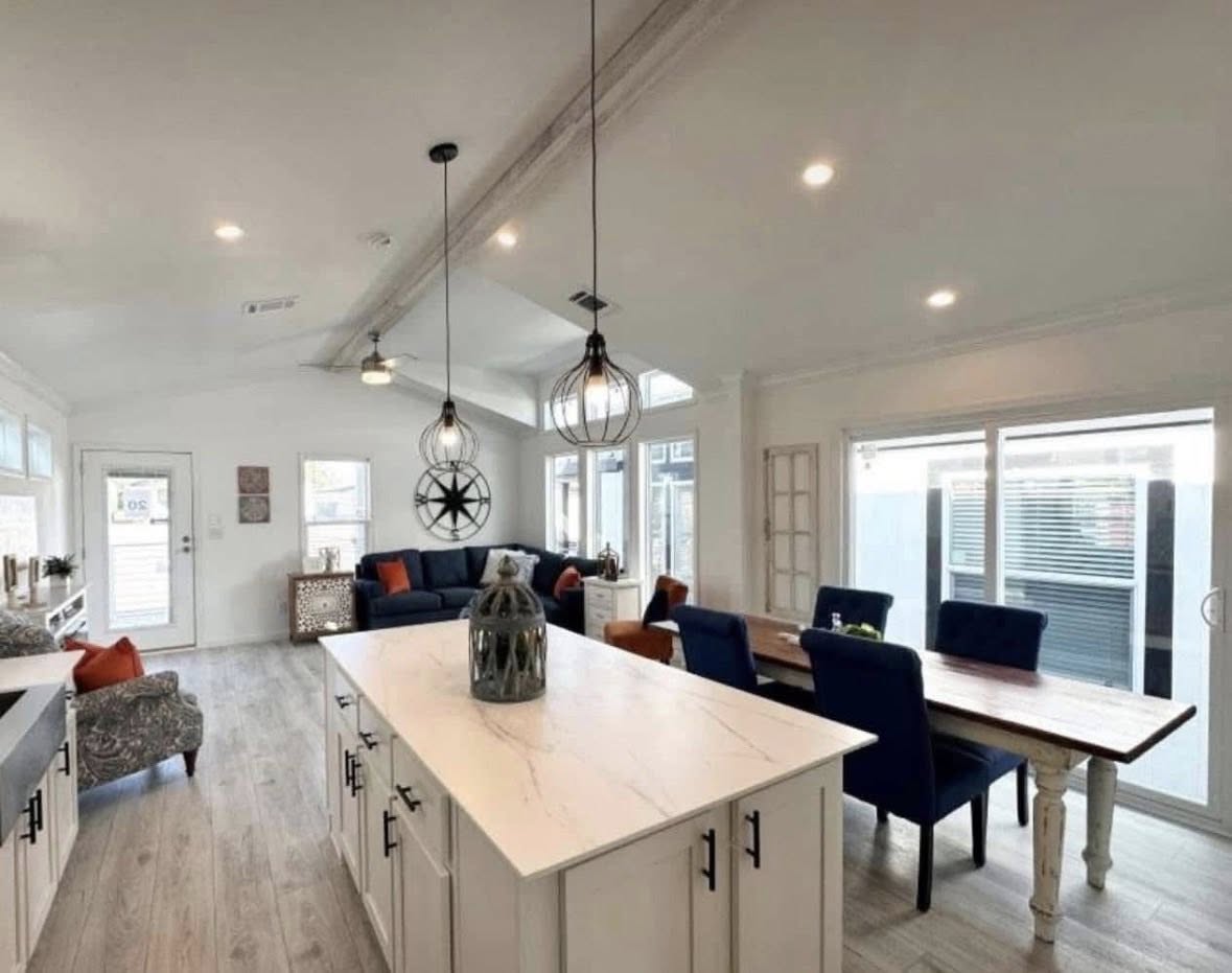 Spacious open-plan living area with a white kitchen island, blue chairs around a wooden dining table, cozy sofas, and modern pendant lights, exuding a warm, inviting ambiance.
