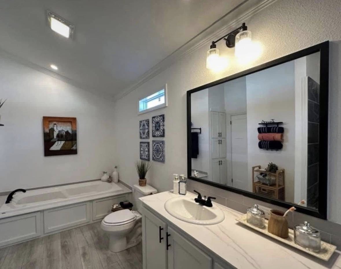 A modern bathroom with white walls and wood flooring. It features a large mirror, sink, bathtub, wall art, and towels, conveying a clean and cozy ambiance.