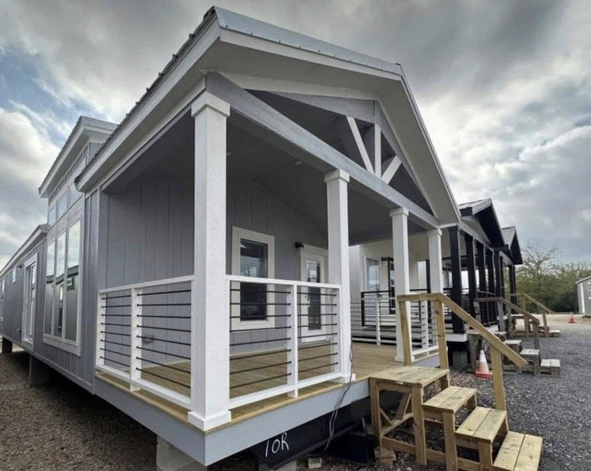 A modern tiny house with a covered front porch, white railings, and light blue siding under a cloudy sky. A gravel path leads to other tiny homes.