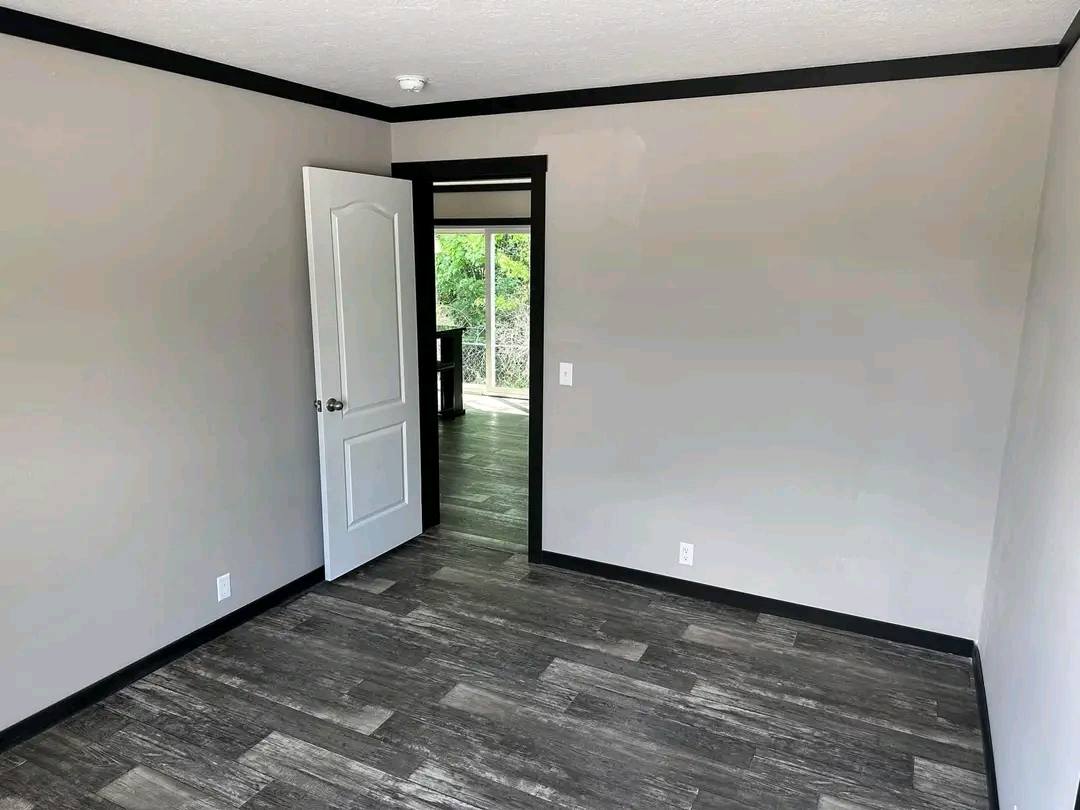 Empty room with gray walls, dark wood flooring, and black trim. An open door leads to a bright adjacent space with visible greenery outside.