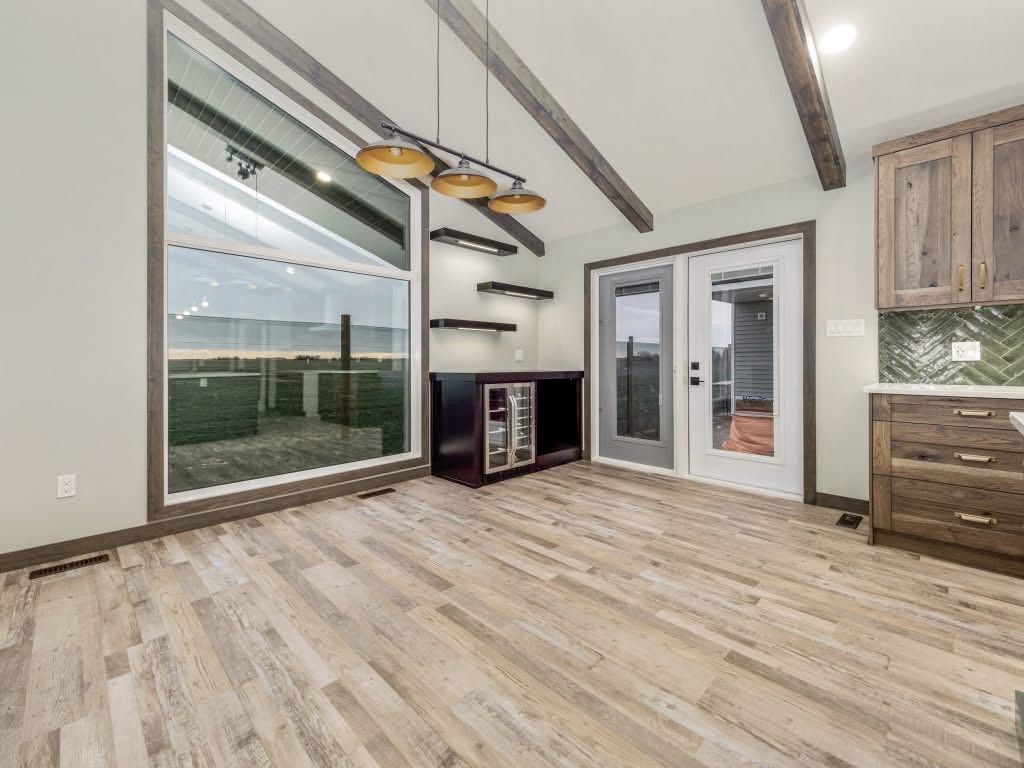 Spacious room with large windows, light wood flooring, and exposed beams. Modern glass double doors, wall shelves, and warm ambient lighting convey a cozy vibe.