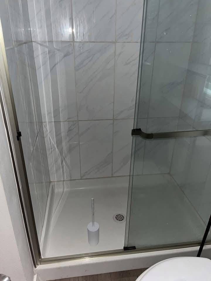 Glass-enclosed shower with white marble tile walls, featuring a handheld showerhead and a cylindrical brush in the corner, conveys a clean, modern bathroom feel.