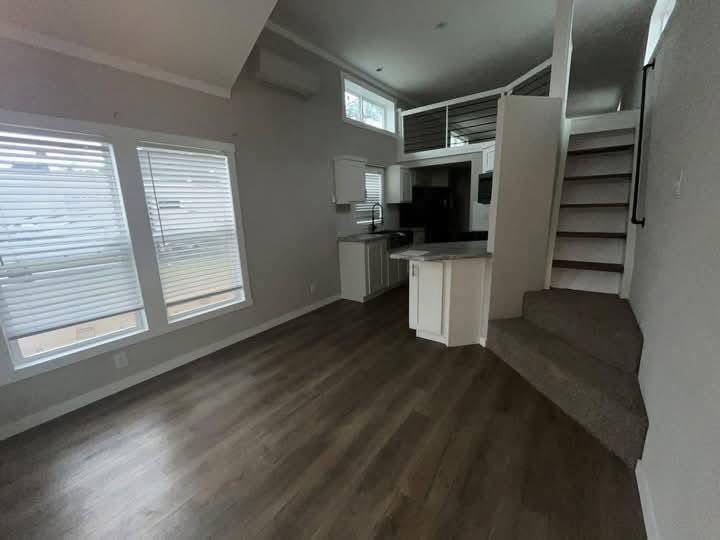 Alt text: A compact, modern living space with a loft design. The room features dark wood flooring, a small kitchen area with white cabinetry, and a staircase leading to an elevated loft. Large windows on the left allow natural light, creating a cozy atmosphere.