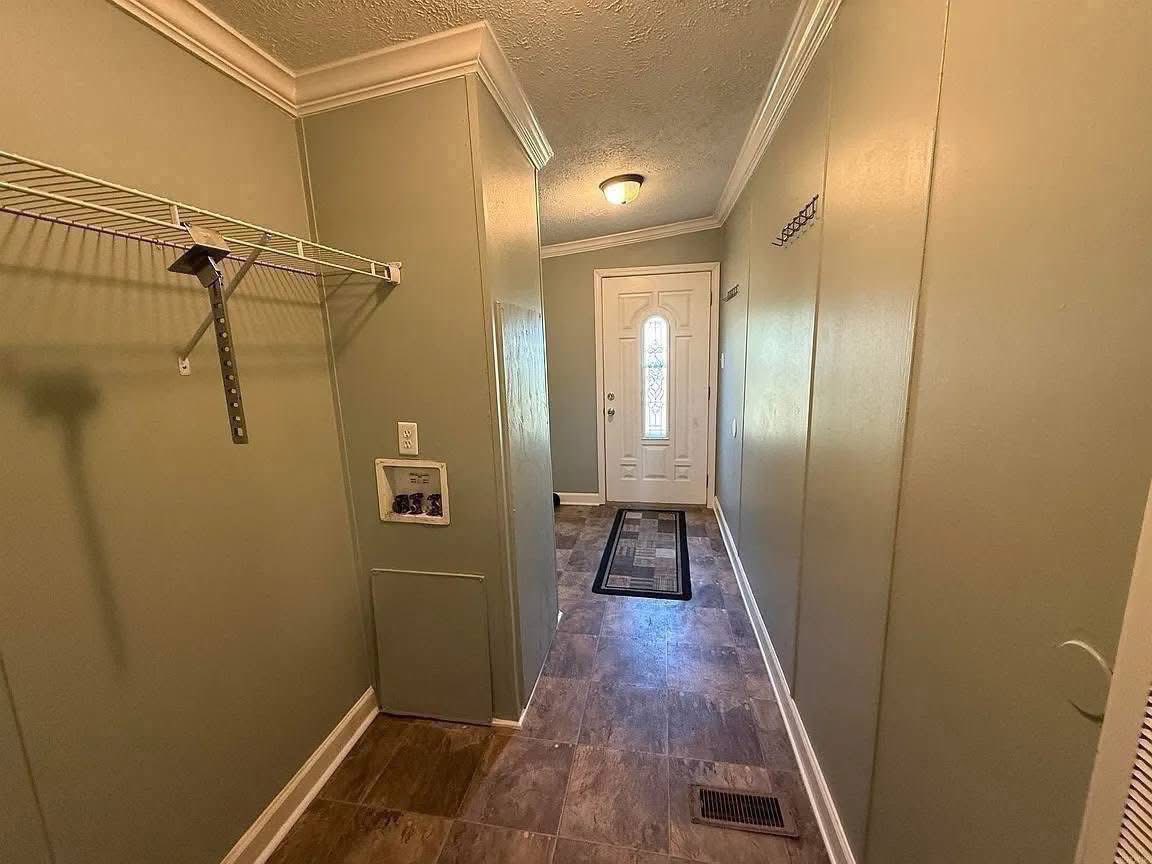 A narrow hallway with light green walls and tiled floor leads to a white front door with a window. A ceiling light brightens the space. A wire shelf is on the left, and a small patterned rug lies in front of the door.