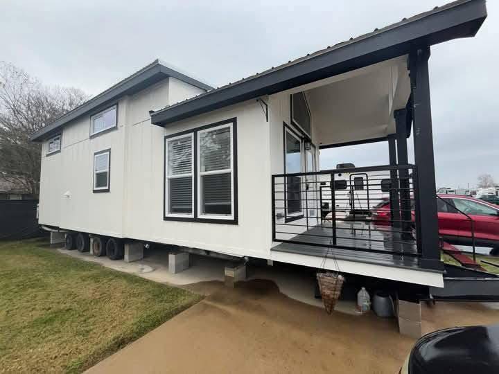 Modern small house with white siding and black trim, elevated on a concrete base. It features large windows, a covered porch, and a lawn under an overcast sky.