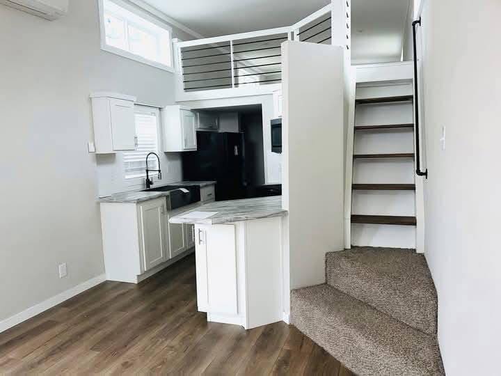 Modern open-concept tiny home interior with white cabinets, dark countertops, and wood flooring. A staircase leads to a loft area, creating a cozy atmosphere.