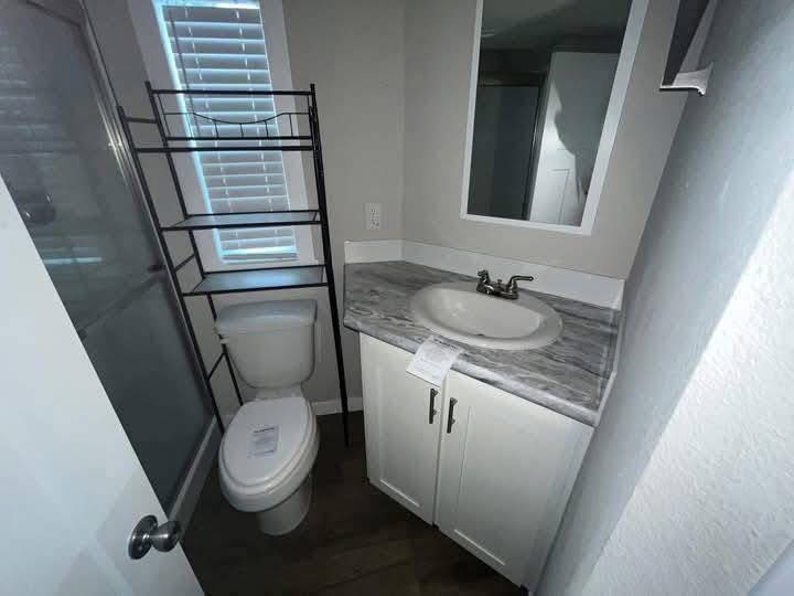 Compact bathroom with modern gray and white decor. Features a small sink with marble countertop, toilet beside a towel rack, and a narrow window.