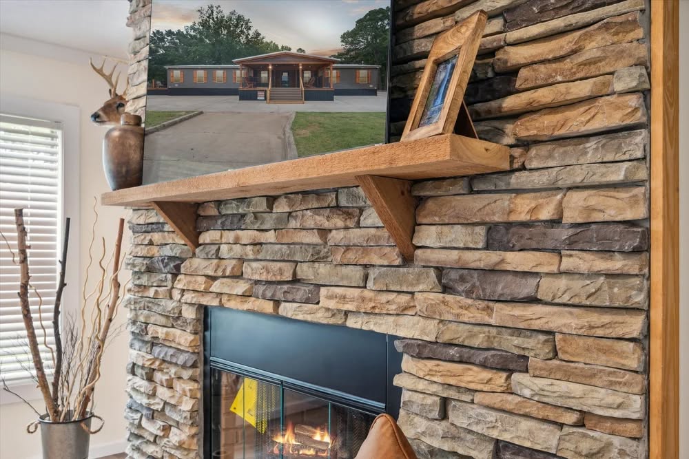 Cozy stone fireplace with burning logs, wooden mantle displaying a picture frame and decorative vase. Warm, inviting ambiance with rustic elements.