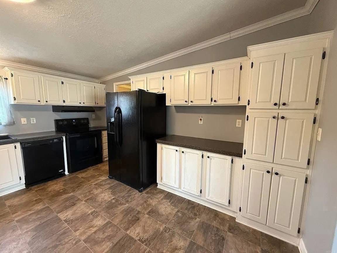 A modern kitchen features white cabinetry, dark countertops, and a black refrigerator. The space is open and bright with tiled flooring and gray walls.