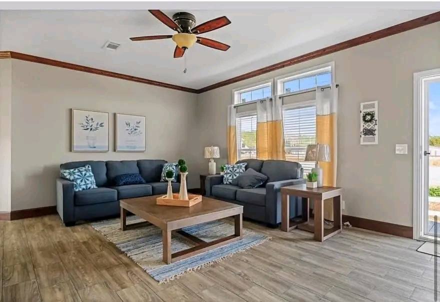 A cozy living room with two gray sofas, blue and patterned cushions, a wooden coffee table, potted plants, and large windows with yellow curtains.