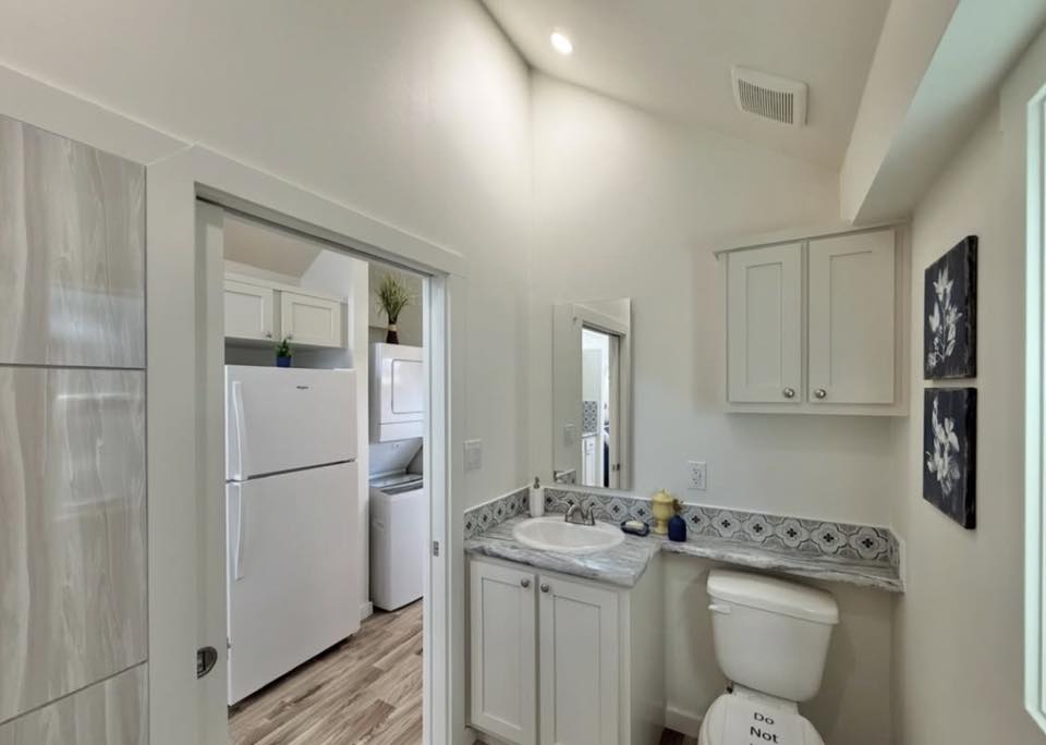 A brightly lit bathroom with white cabinets, marble countertops, and a patterned backsplash. A door opens to a laundry area with stacked appliances.