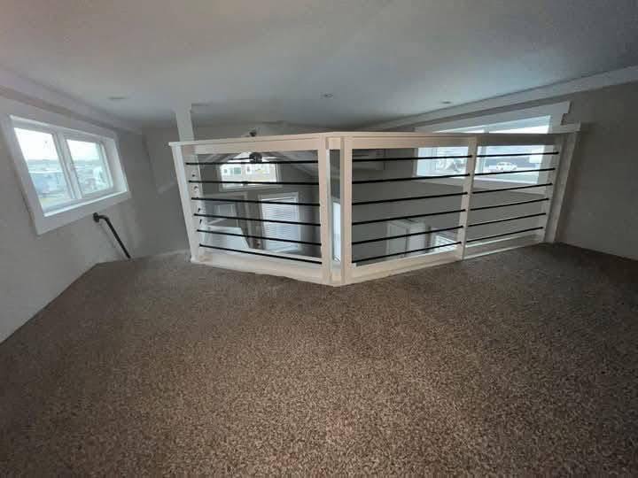 A loft space with beige carpet and a white railing overlooks a lower floor. Natural light streams in from two windows, creating an airy feel.