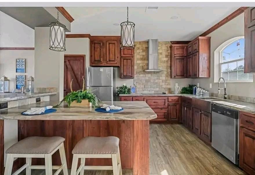Warm kitchen with wooden cabinets, stone island, and pendant lights. Cozy stools and plants add charm. Sunlit atmosphere with modern appliances.