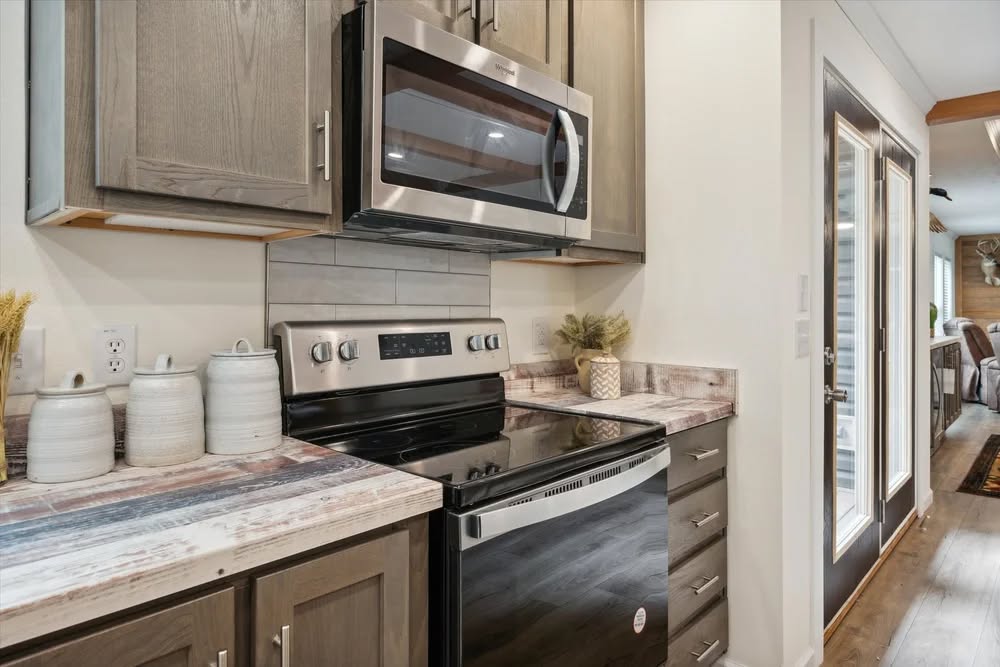 Modern kitchen corner with a stainless steel stove and microwave. Wooden cabinets, rustic countertop, ceramic containers, and potted plant enhance warmth.