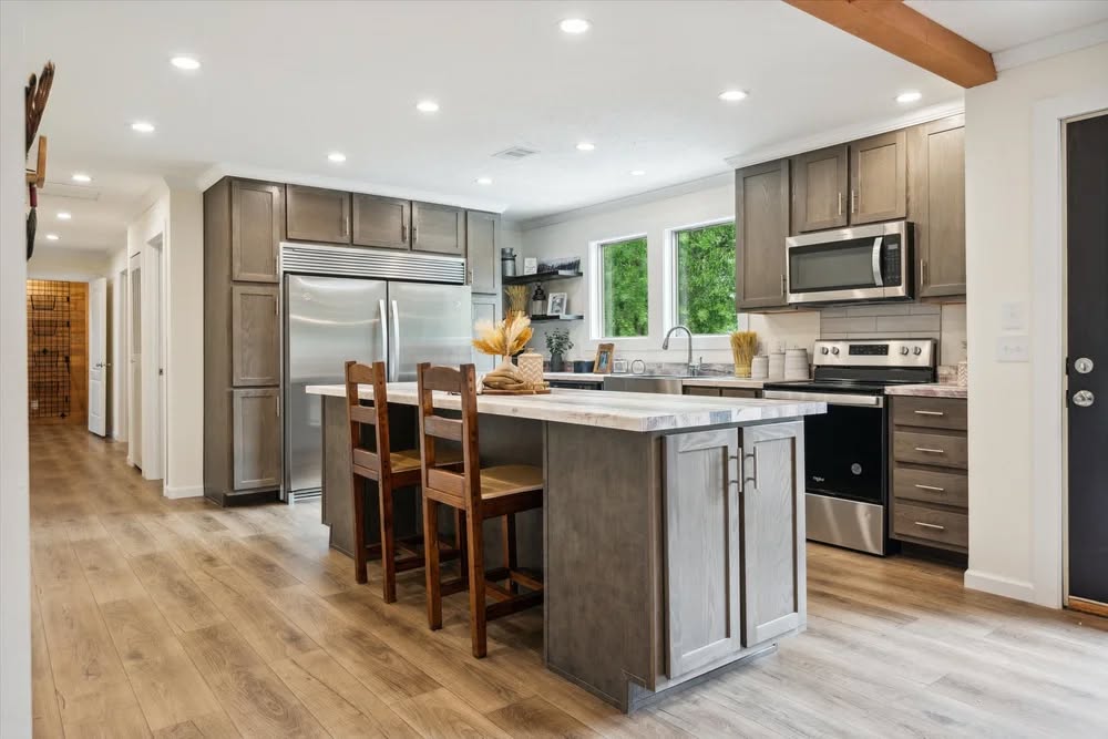Spacious modern kitchen with wood cabinets, large island, and bar stools. Features stainless steel appliances and bright windows with lush greenery.