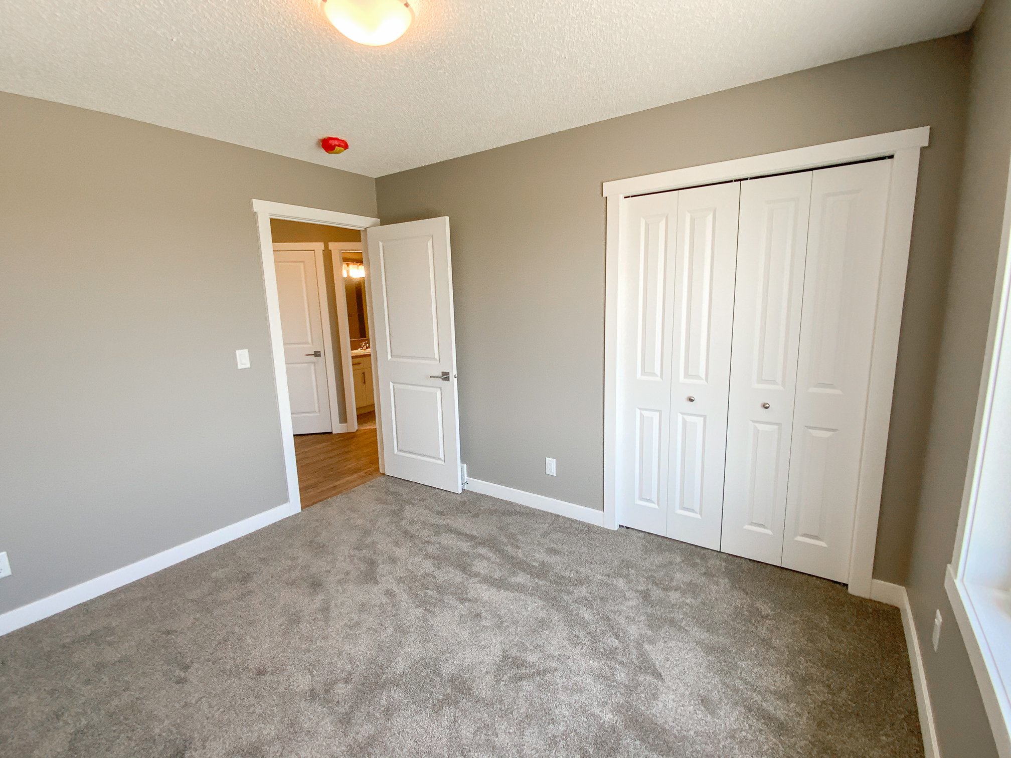 Empty room with beige walls, gray carpet, and white trim. An open door leads to a lit hallway. White folding closet doors are on the right wall.