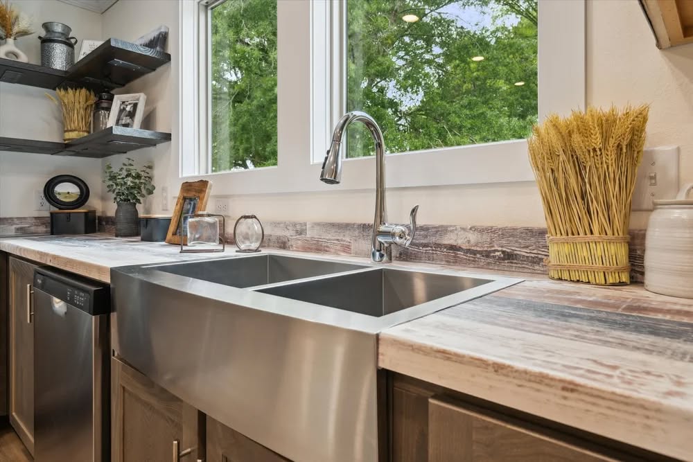 Modern kitchen with large stainless steel sink, wood countertops, and open shelves. A window shows lush greenery outside, creating a tranquil ambiance.