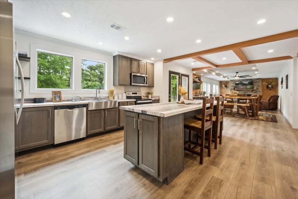 Spacious kitchen with gray cabinets, stainless steel appliances, and wooden island. Open to a dining area with wooden beams and cozy decor. Bright, inviting ambiance.