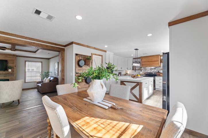 Wooden dining table with white chairs in a bright, open plan kitchen and living room. Green plant centerpiece, rustic decor, modern, warm atmosphere.