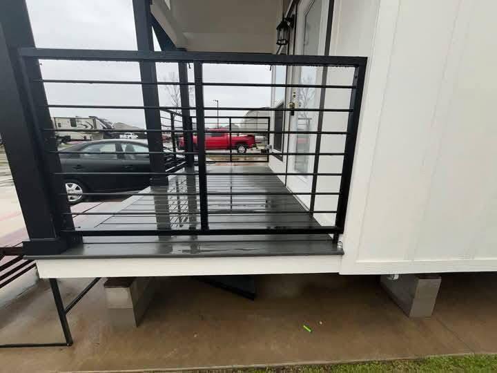 A small porch with black metal railing attached to a white building. Wet concrete floor, with cars and RVs parked outside on an overcast day.