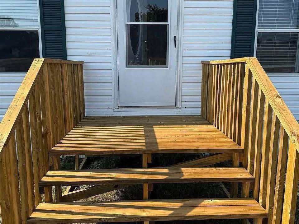 Wooden porch with railing and stairs leading to a white door on a house with white siding. Sunlight casts shadows, creating a warm, inviting tone.