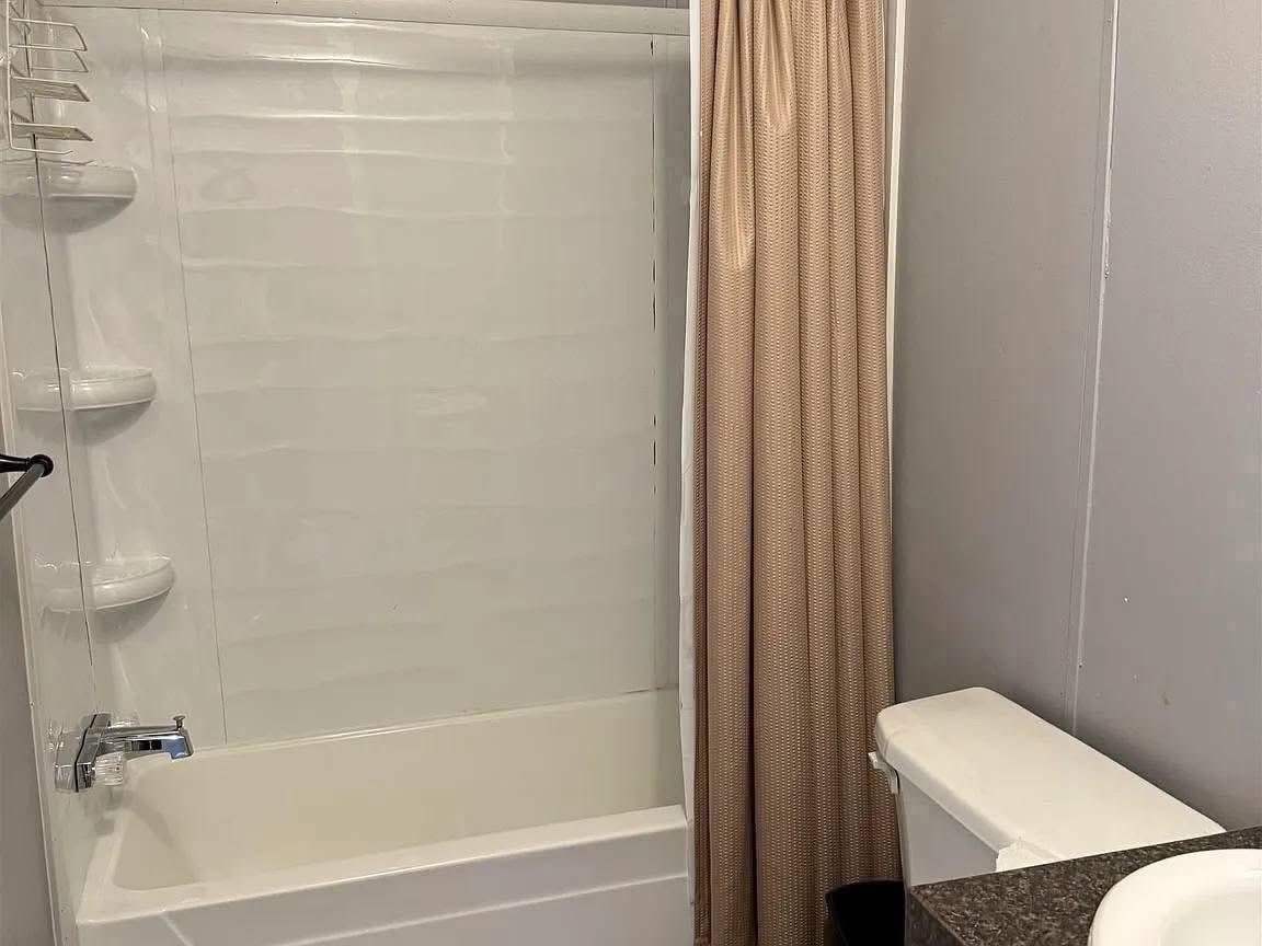 A clean bathroom features a white bathtub with a beige shower curtain partially closed. The room includes a countertop with a glimpse of a white sink.
