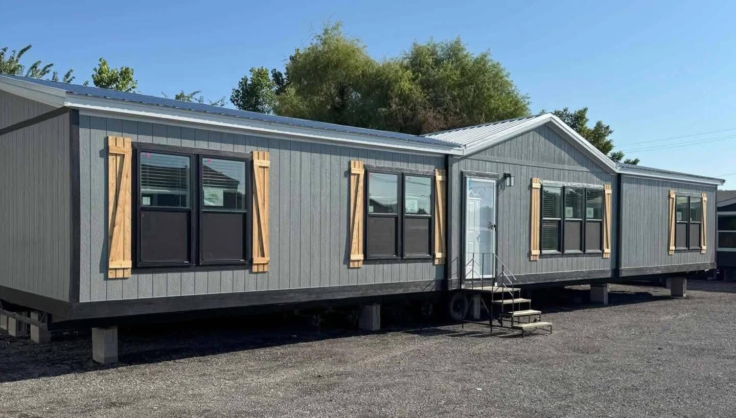 A gray mobile home with wooden shutters and large windows, set on a gravel lot. Small steps lead to the door, with trees visible in the background.