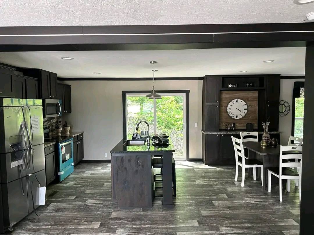 Modern kitchen with dark wood cabinets, stainless steel appliances, and a central island under pendant lighting. A dining area with white chairs and a large clock on the wall adds contrast. Bright daylight enters through a sliding glass door, creating a welcoming atmosphere.