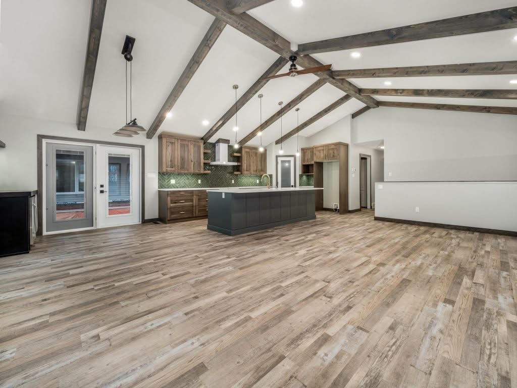 Spacious modern kitchen with vaulted ceiling and exposed wooden beams. Features a dark island, pendant lights, wooden cabinets, and large glass doors.