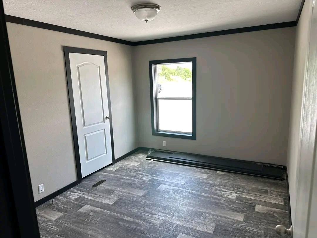 Small empty room with light gray walls, a white door, and a single large window. Dark gray trim and wood-look flooring create a minimalist, modern feel.