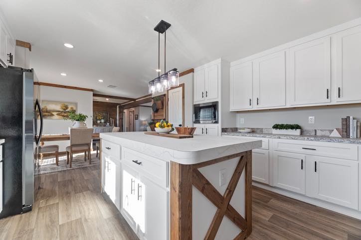 Modern kitchen with white cabinets, a large island with wood accents, and hanging lights. In the distance, a dining table is visible. The space feels bright and inviting.