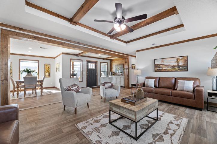 Spacious living room with wooden beams, brown leather sofa, gray armchairs, and a patterned rug. Ceiling fan above and dining area in the background.