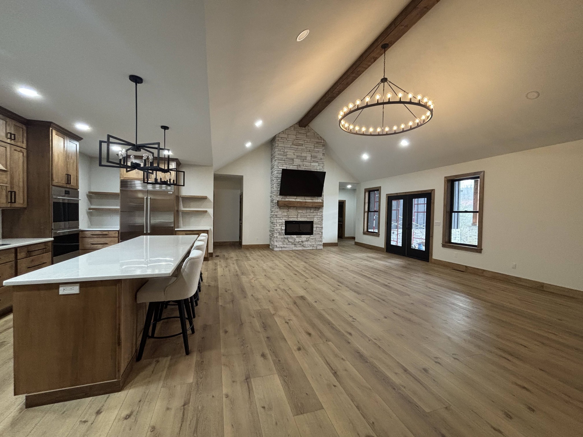 Spacious open-concept room with a modern kitchen on the left, featuring wooden cabinets and a white island. A cozy stone fireplace and large chandelier add warmth.