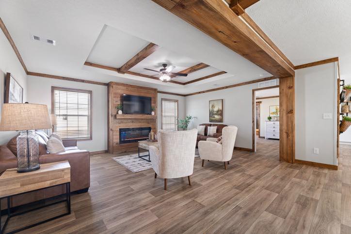 Cozy living room with wooden beams, white walls, and wood flooring. Features a sofa, two cream chairs, a TV above a fireplace, and soft lighting.