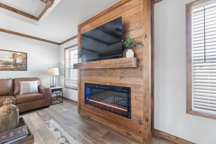 Cozy living room with a mounted TV above a modern fireplace on a wooden accent wall. Brown sofa, lamp, and framed artwork create a warm atmosphere.