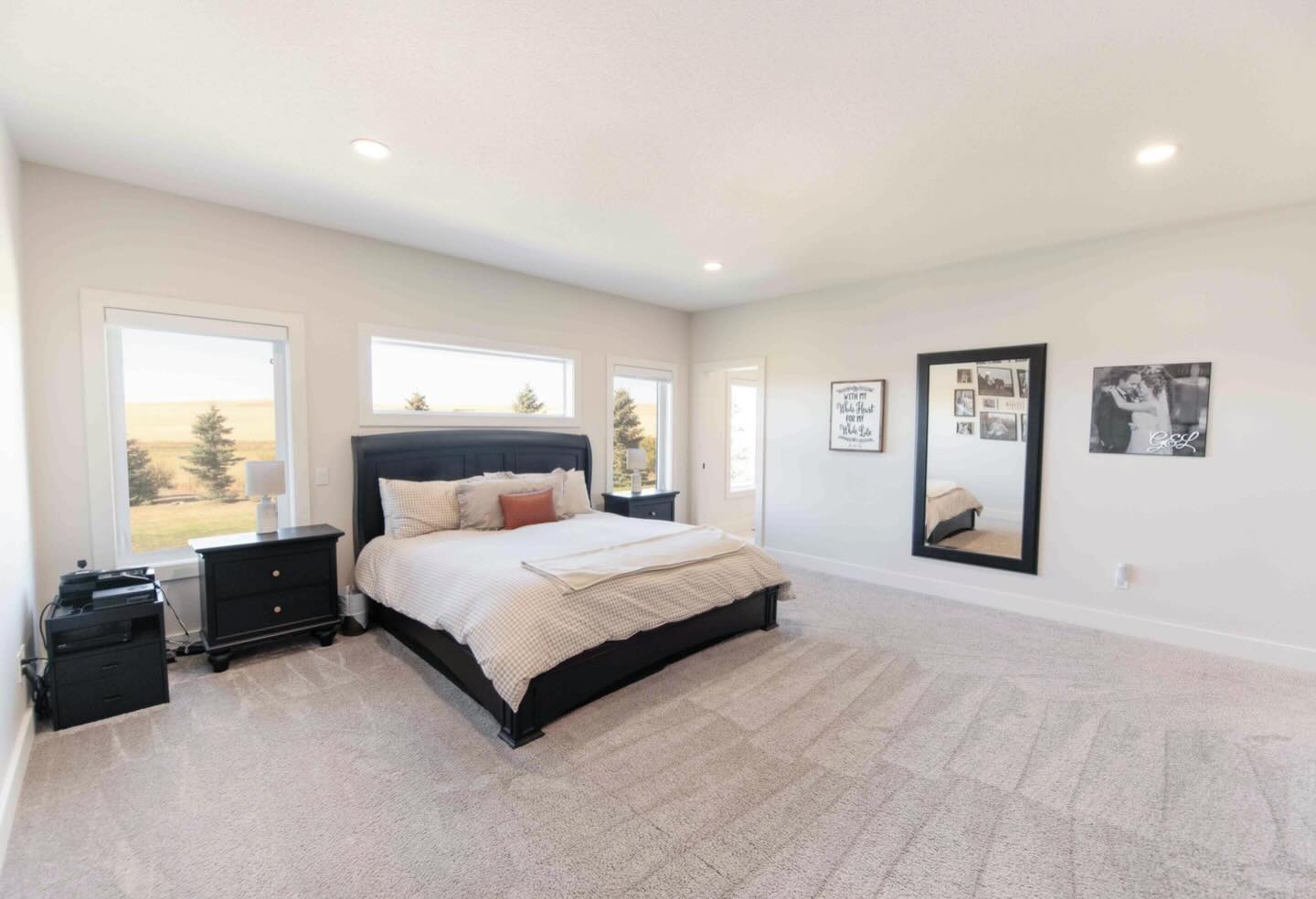 Spacious bedroom with a large bed, black headboard, and soft linens. Two nightstands flank the bed. Wall art and a full-length mirror add warmth.