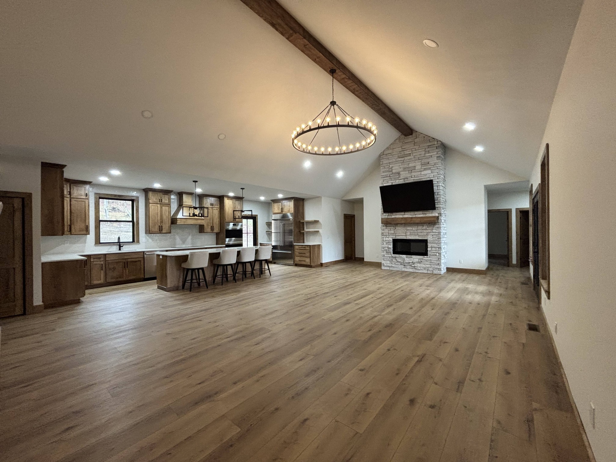 Spacious open-concept room with a modern kitchen, wooden cabinetry, a large island with bar stools, and a stone fireplace under high ceilings. Cozy ambiance.