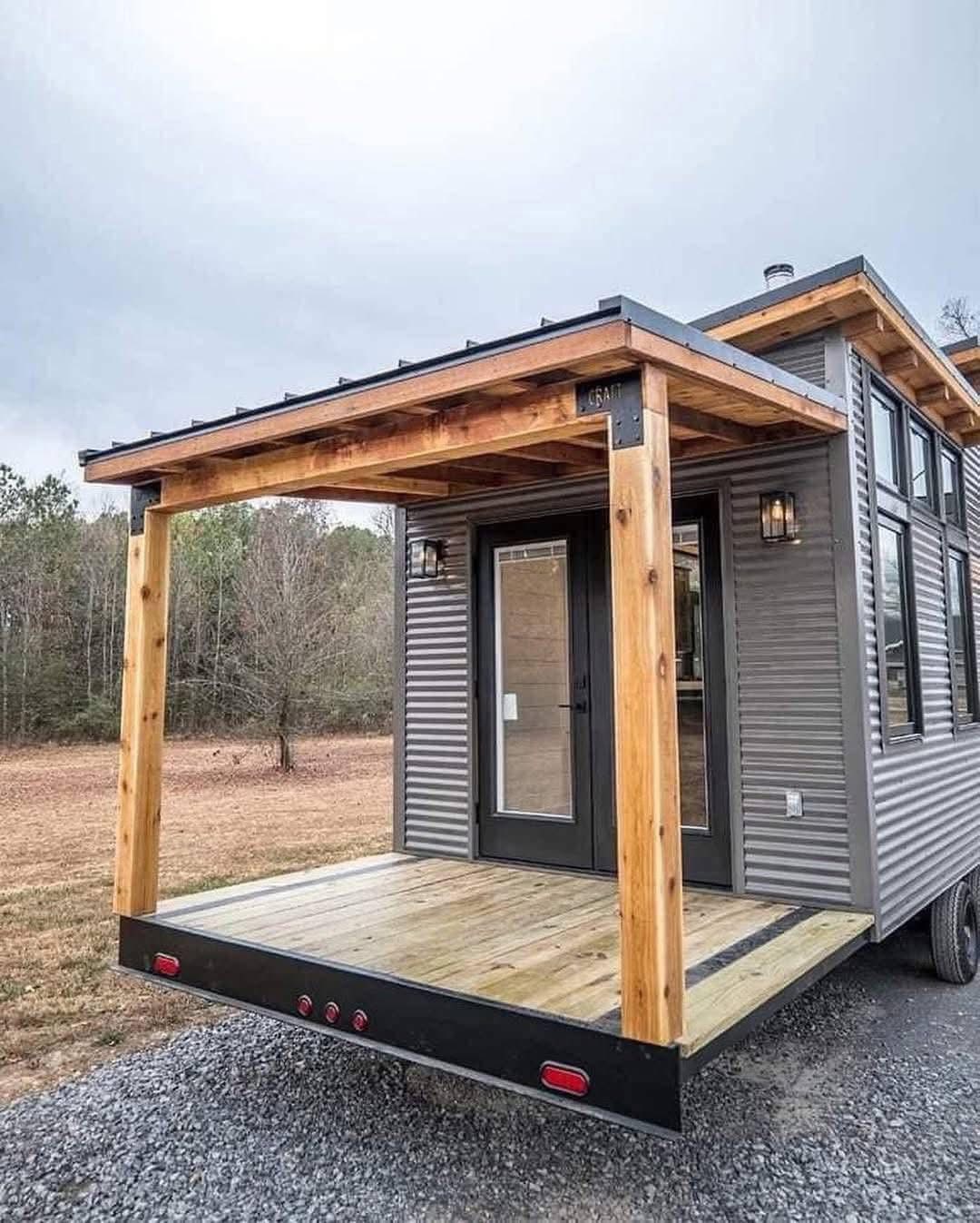 A modern tiny house on wheels with a wooden porch and large glass doors. The corrugated metal siding gives a sleek look against a rural backdrop.