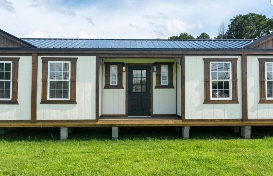 Small modular home with a black metal roof and white siding, accented by brown trim. It stands on a grassy lawn under a partly cloudy sky.