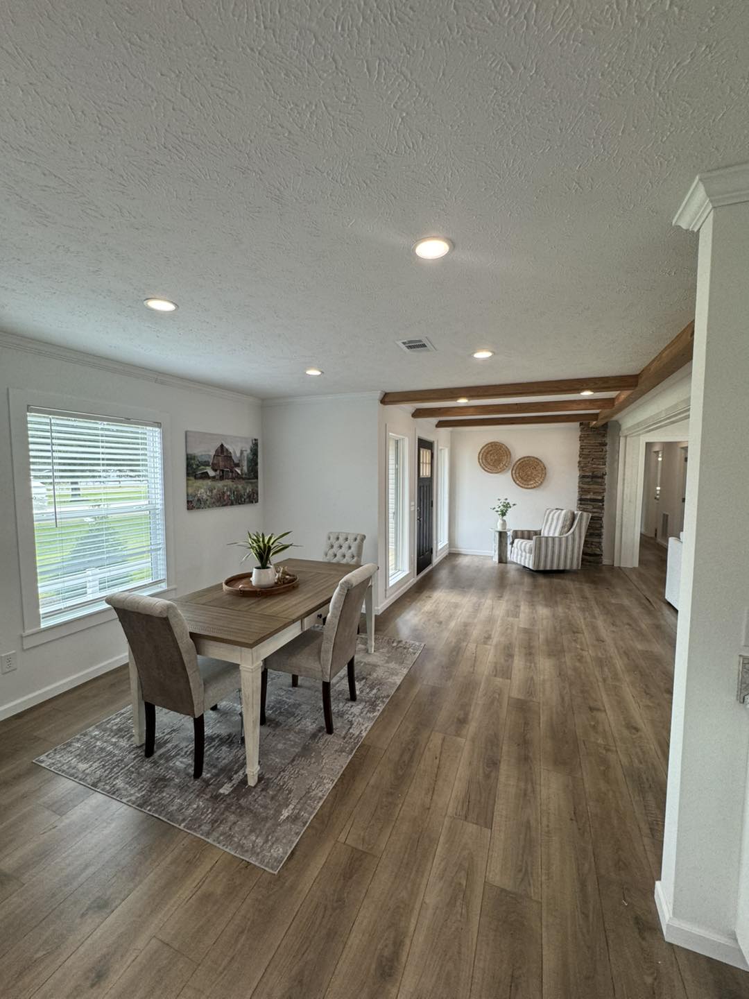 Modern dining and living area with wood flooring, white walls, and ceiling beams. A table with four chairs is on the left, and armchairs with decorative wall elements are in the background, creating a cozy ambiance.