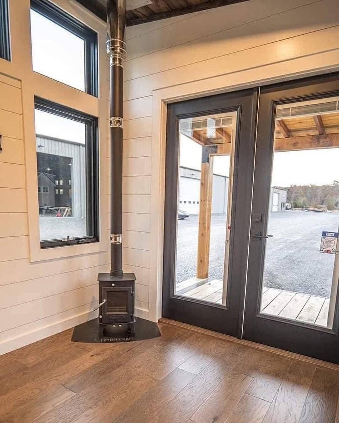 Corner of a cozy room with a small black woodstove near a window and glass door. The room has light wood walls and dark wooden floors.