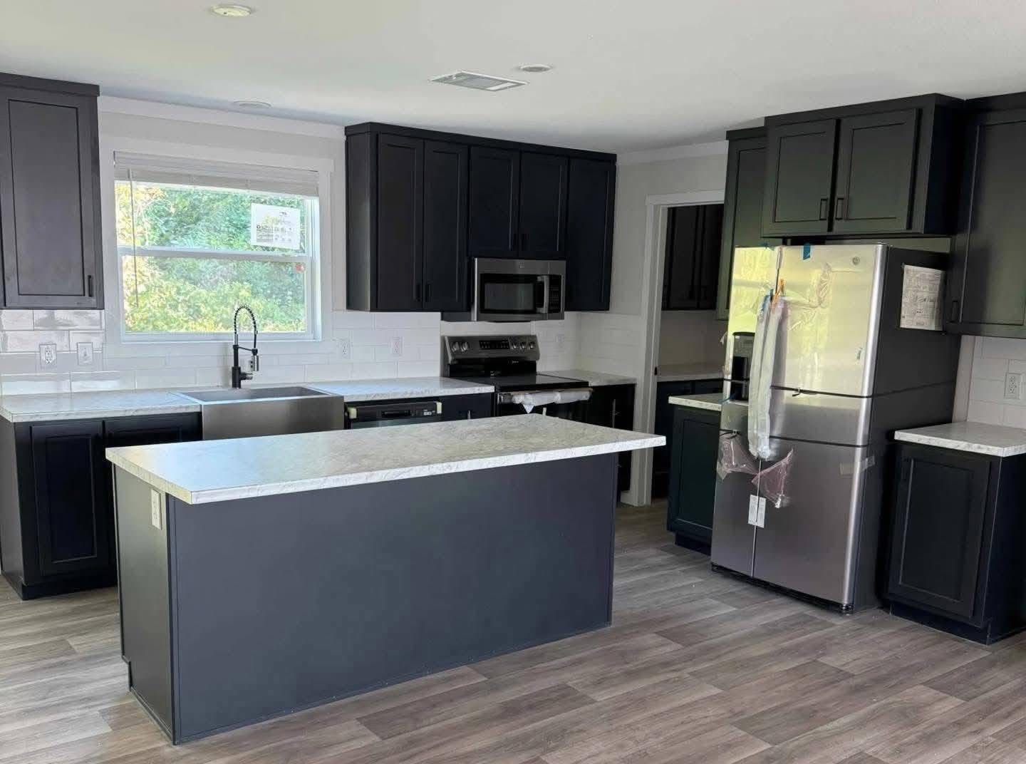 Modern kitchen with sleek black cabinets, stainless steel appliances, and a central island. Light wood flooring and sunlight through a window create a bright atmosphere.