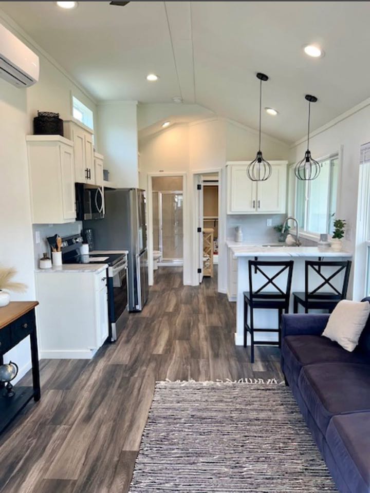 Modern tiny home interior featuring a sleek kitchen with stainless steel appliances, white cabinets, a cozy living room with a blue sofa, and a gray rug.