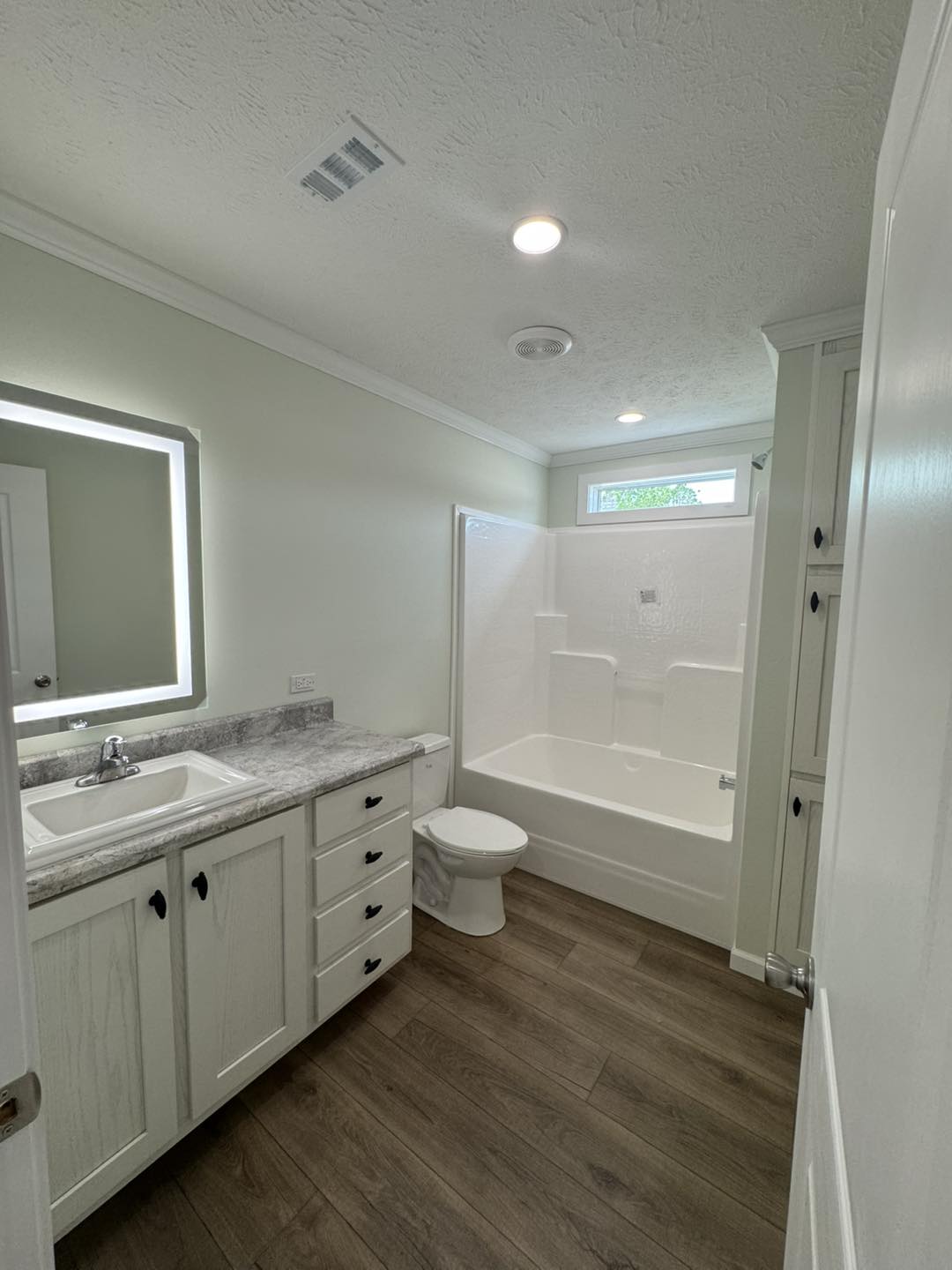 A modern bathroom with wood flooring features a sink with a granite countertop and cabinets on the left, a toilet, and a bathtub on the right under a small window.