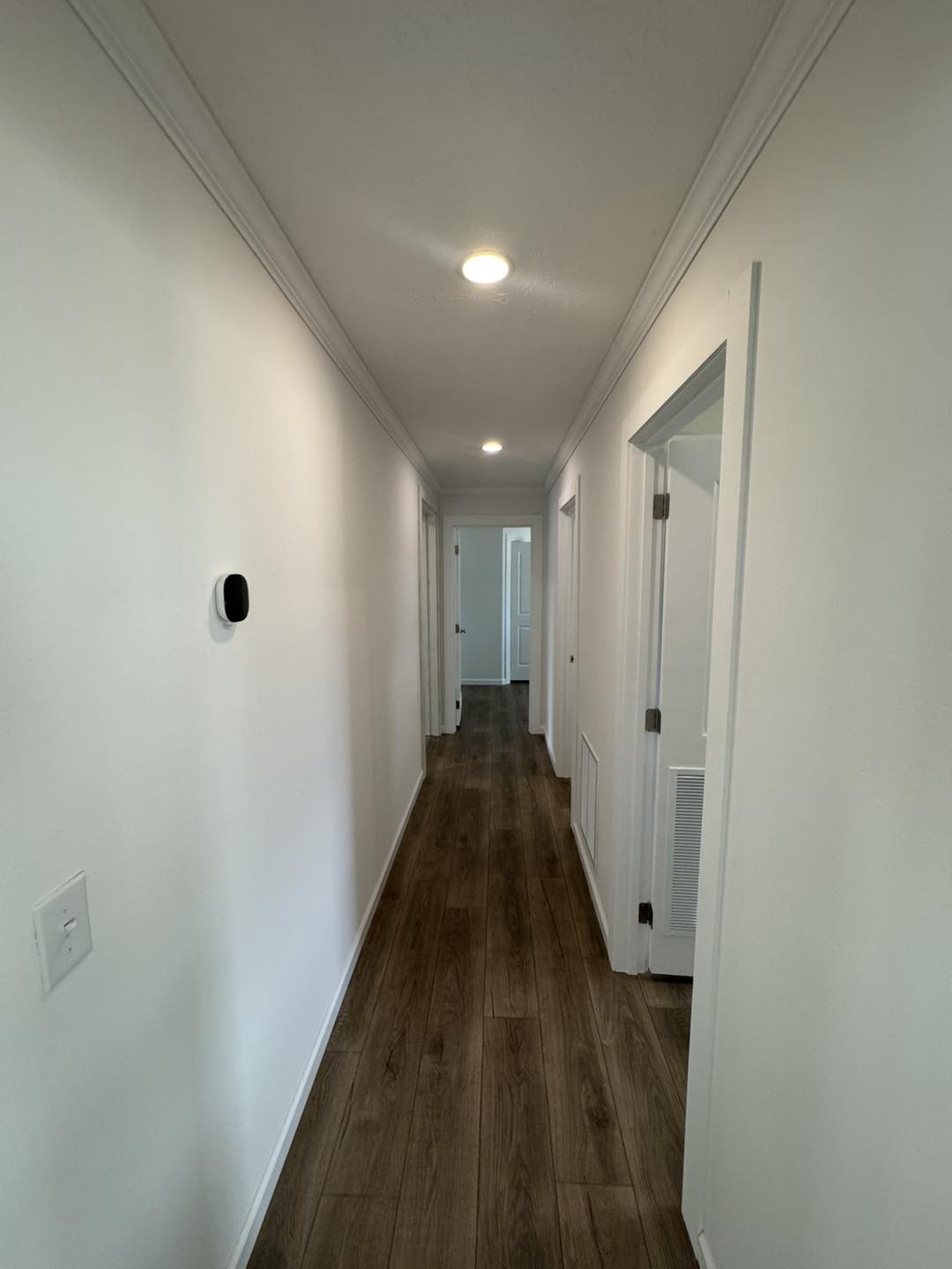 A narrow, well-lit hallway with white walls and dark wooden flooring. Recessed ceiling lights illuminate the space, with doors on the right side.