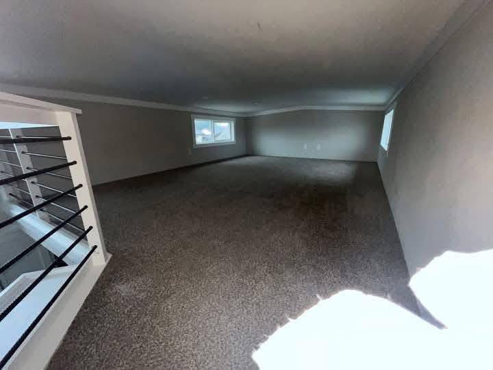 A small, carpeted attic room with sloped ceilings is lit by two windows. It has a white railing with black bars on the left, creating a cozy atmosphere.